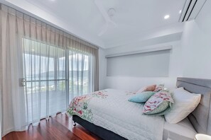 A bedroom with a bed, a large window with curtains, a ceiling fan, and a wall-mounted air conditioning unit. - The View (Airlie Beach)