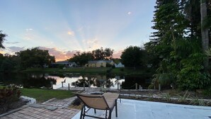 Eine Terrasse mit einem Stuhl mit Blick auf einen See, umgeben von viel Grün und einem Haus im Hintergrund.