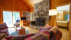 A cozy cabin interior with a stone fireplace, a television, a bookshelf, and plaid furnishings.