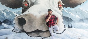 Une personne posant devant une grande fresque murale représentant le visage d’une vache, avec une montagne enneigée en arrière-plan.