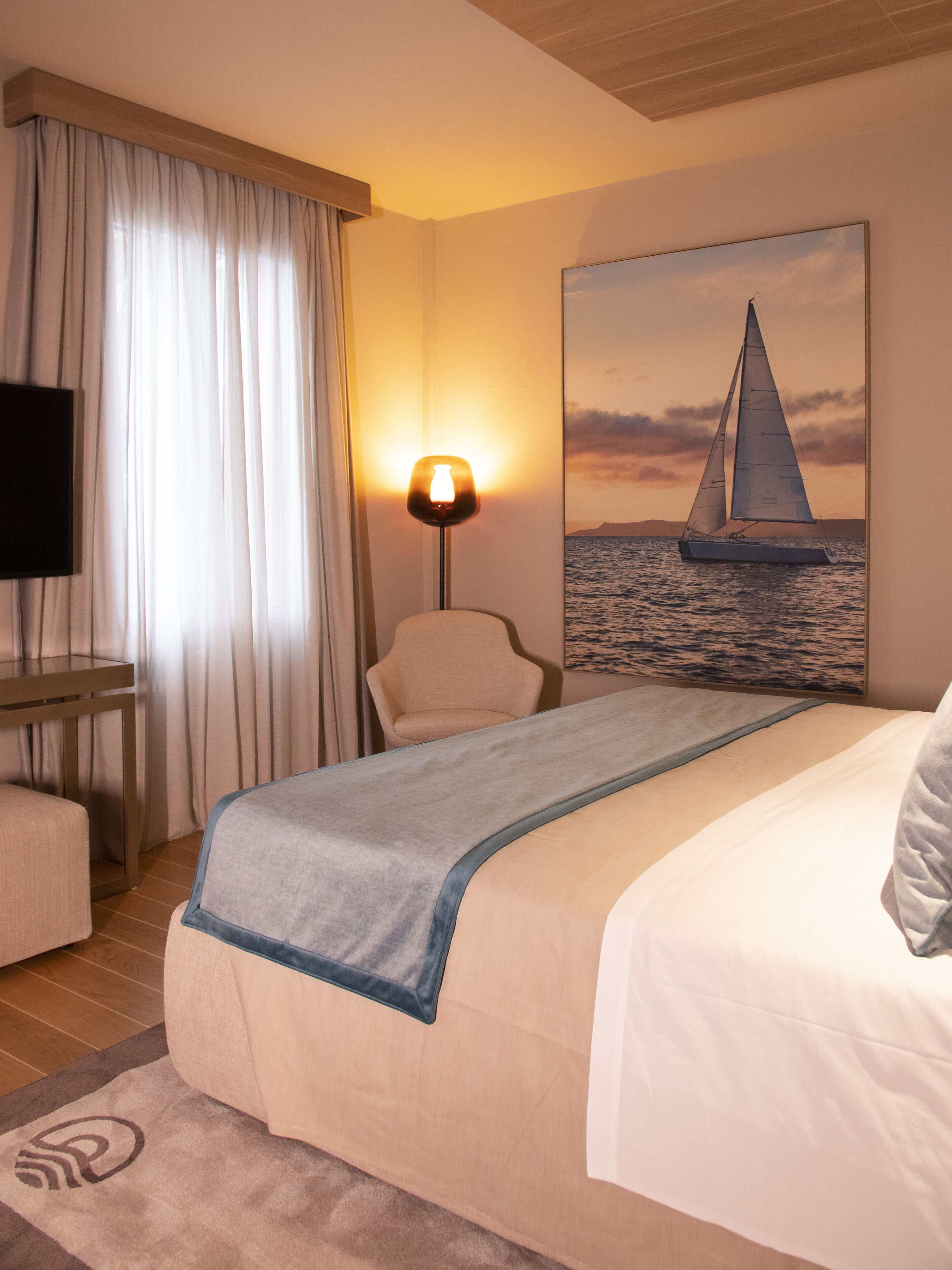 Habitación de hotel con cama, una silla, un escritorio, una lámpara y un cuadro de un velero en la pared.