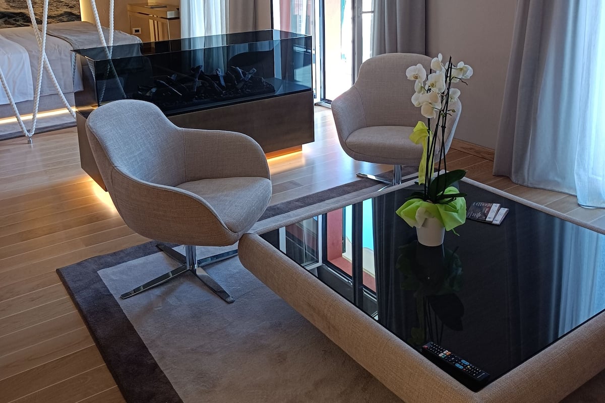 A modern living room with a glass coffee table, two armchairs, a TV, and a view of the city.