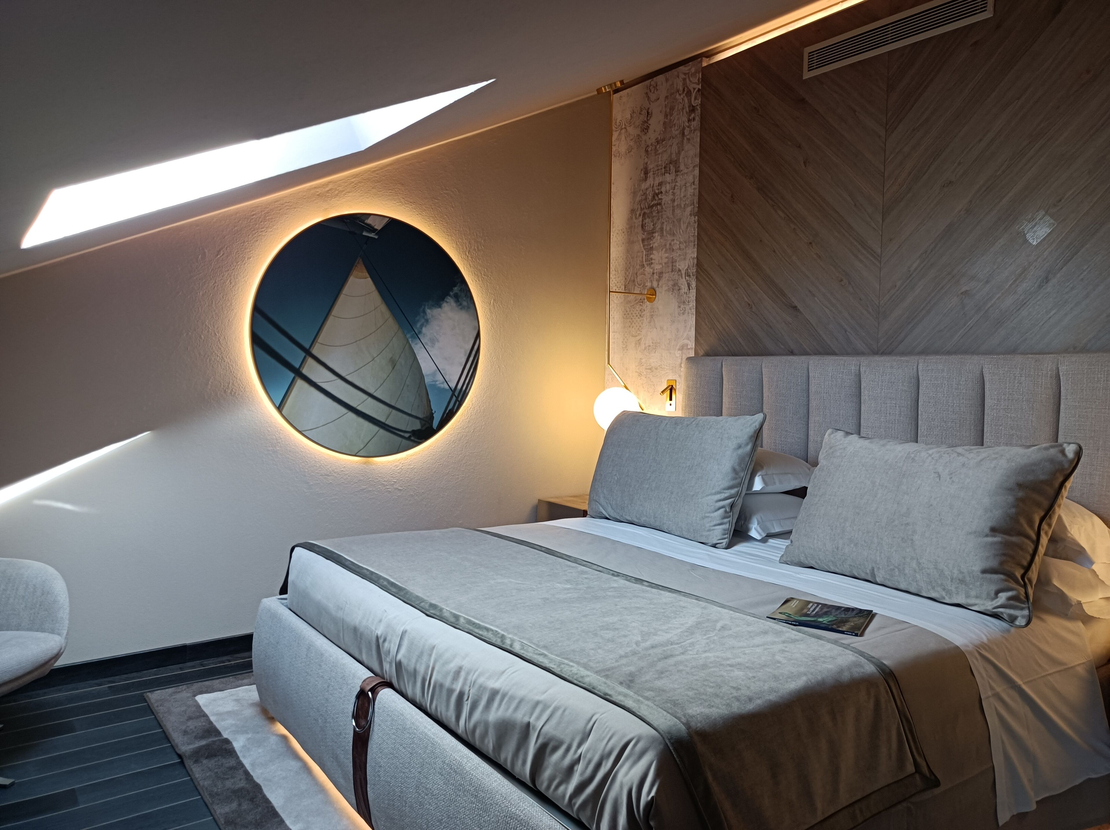 Un dormitorio moderno con tragaluz, un espejo redondo en la pared y una cama con muchas almohadas.