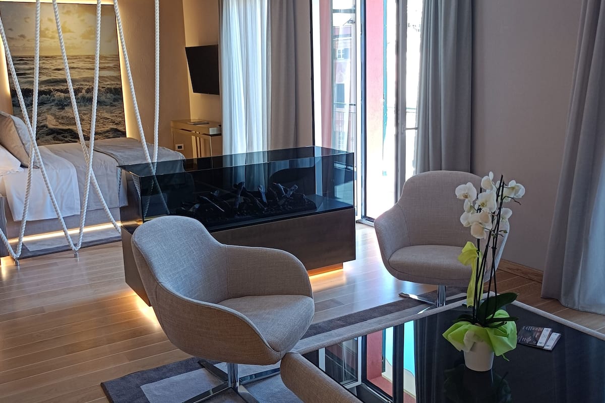 A modern hotel room with a glass coffee table, two armchairs, a sofa, a TV, and a large window with curtains.
