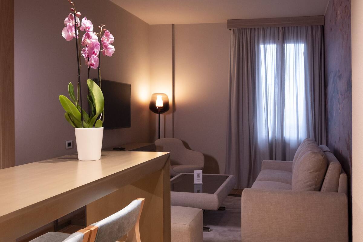 A modern living room with a wooden dining table, grey upholstered chairs, a beige sofa, and a potted orchid plant.