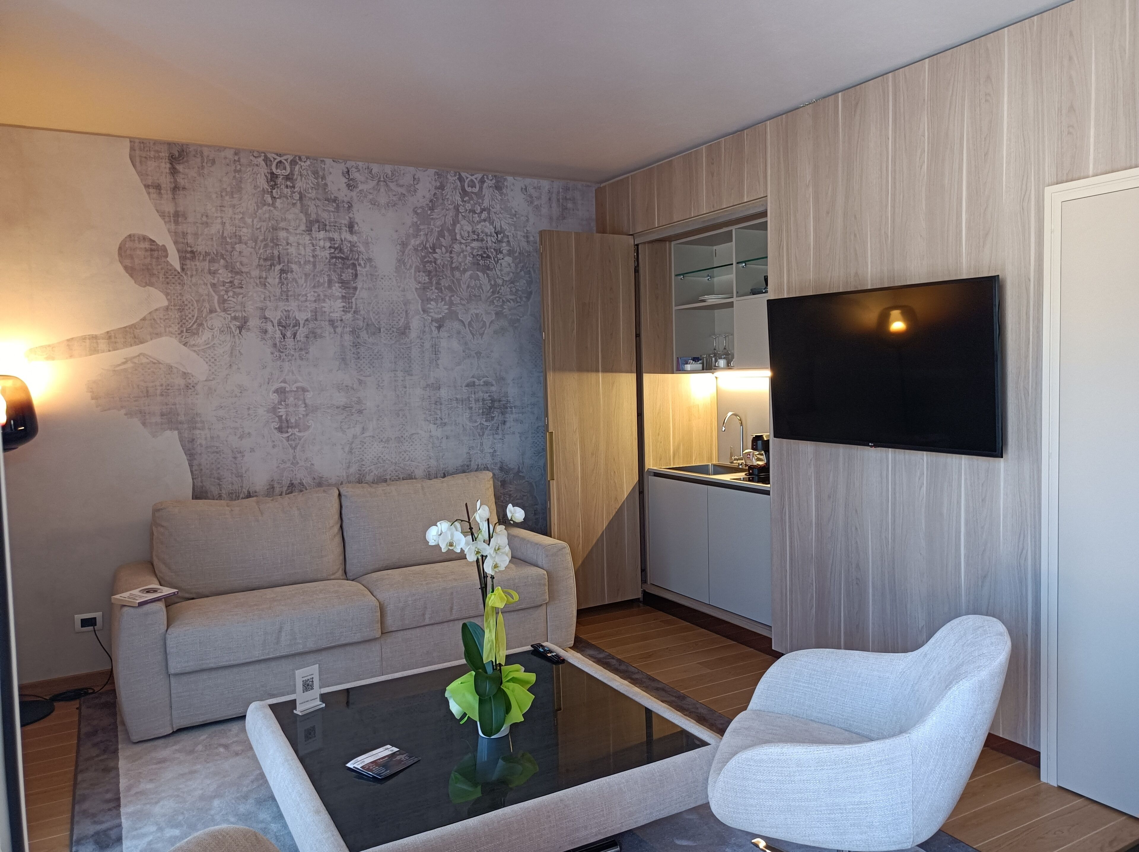 Una sala moderna con un sofá, una mesa de centro con un florero, un televisor de pantalla plana montado en la pared y una cocina con un lavamanos y armarios.