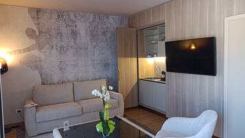 Una sala moderna con un sofá, una mesa de centro con un florero, un televisor de pantalla plana montado en la pared y una cocina con un lavamanos y armarios.
