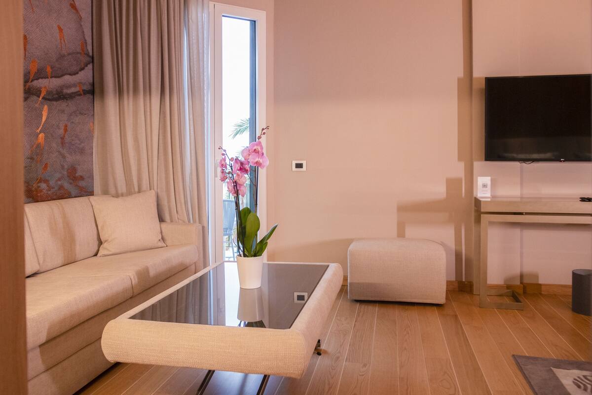 A modern living room with a sofa, a glass coffee table, a small ottoman, a flat-screen TV, and a potted orchid.