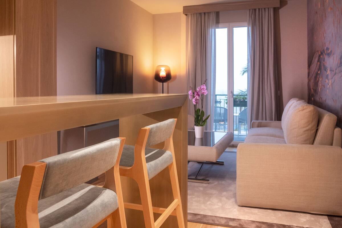 A modern hotel room with a wooden bar, two grey upholstered chairs, a beige sofa, a flat-screen TV, a potted orchid, and a view of the outdoors through large windows.