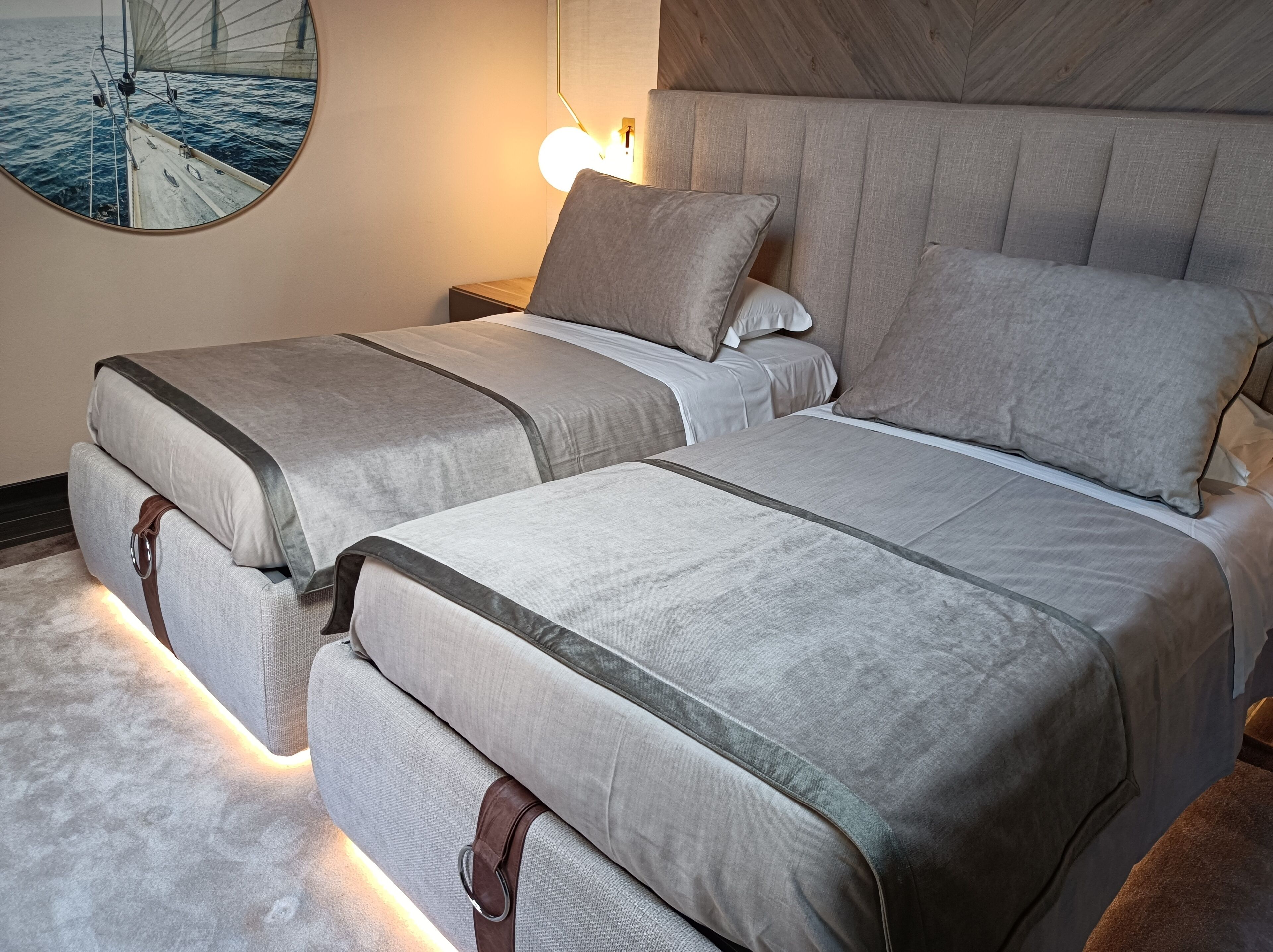 Een tweepersoonsbed met grijze lakens en een hoofdbord, een nachtkastje met een lamp, en een ronde spiegel aan de muur.