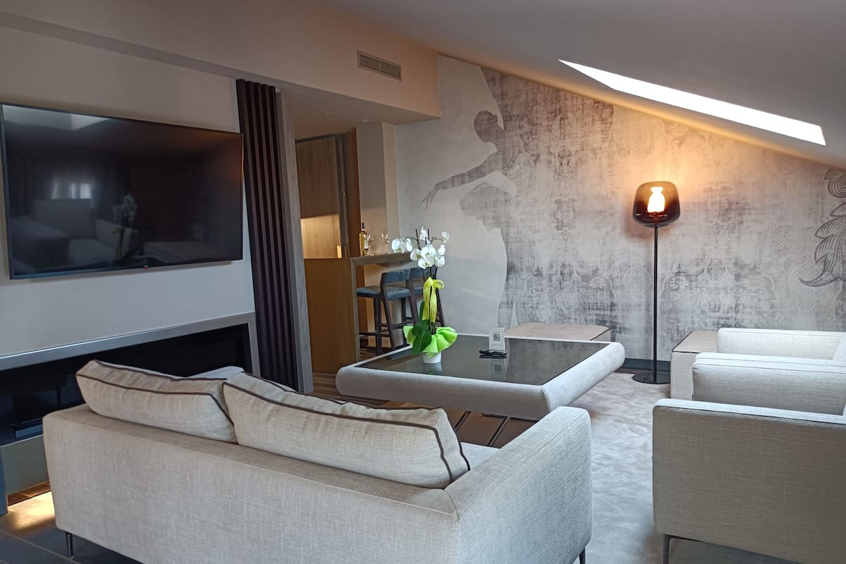 A modern living room with a sofa, a coffee table, and a TV. There is a dining area with a table and chairs. The room has a patterned wall and a floor lamp.