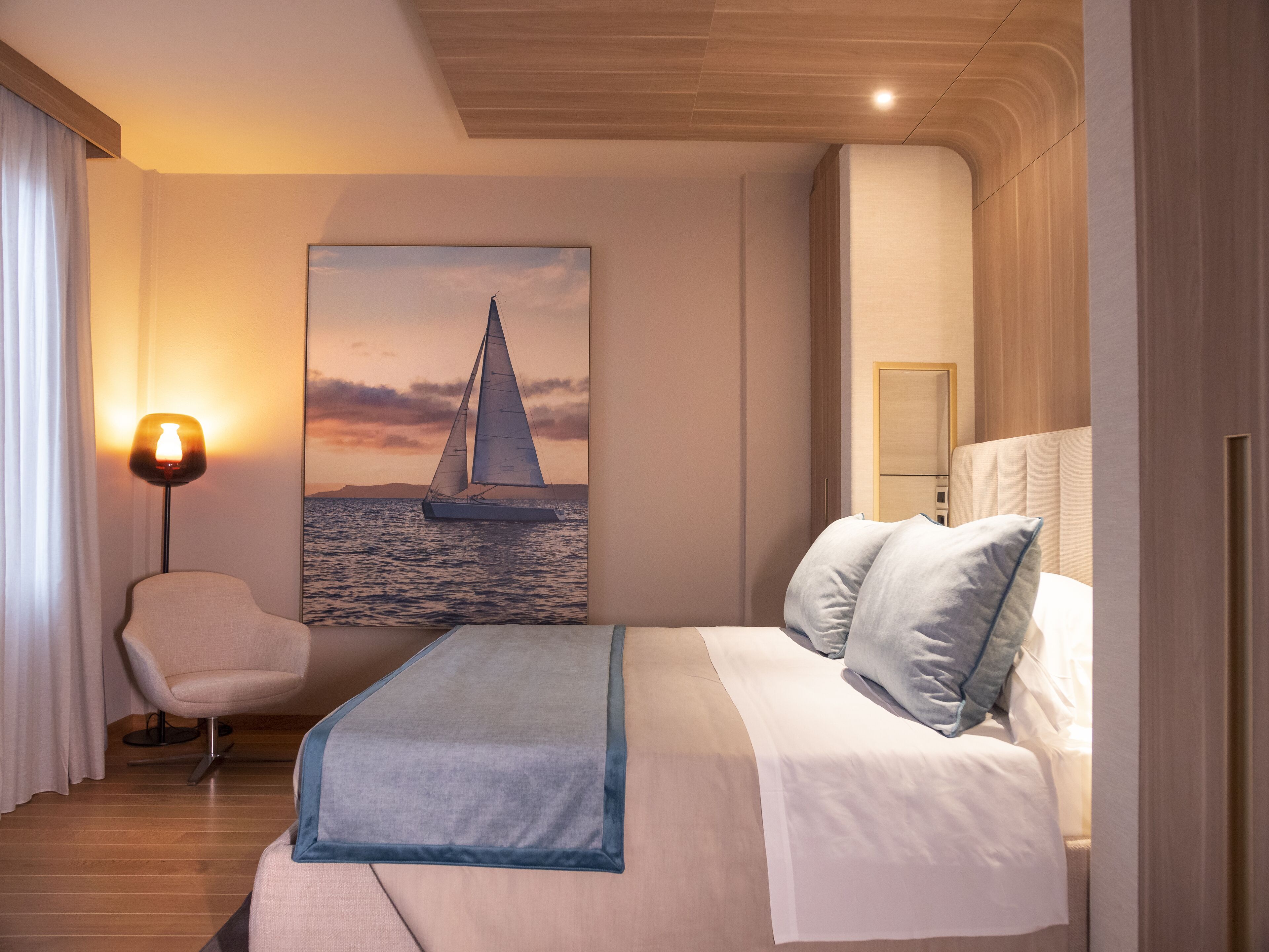 A modern hotel room with a bed, a chair, a painting of a sailboat, and a lamp.