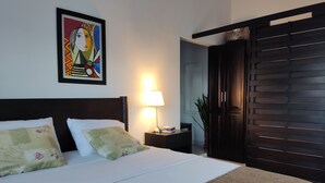 A neatly made bed with white linens and two pillows, a bedside table with a lamp, a framed artwork on the wall, and a dark wooden door. - Casa Ines Hostal a place especially for you... (Santa Marta)