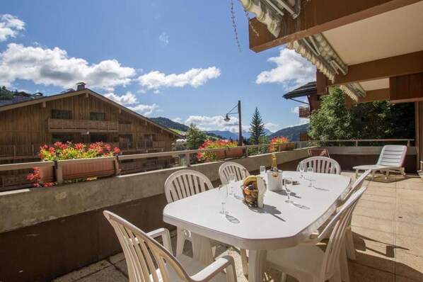 An outdoor patio with a table set for dining, surrounded by white chairs, overlooking a wooden building and mountains.