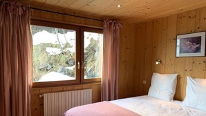 A bedroom with a wooden paneled ceiling, a bed with white linens, a window with curtains, and a radiator below the window. - 100m from resort center, 1st floor, view mountain/street/ski slopes, terrace, fireplace or stove (Le Grand-Bornand)