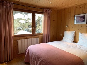 A bedroom with a wooden paneled ceiling, a bed with white linens, a window with curtains, and a radiator below the window.