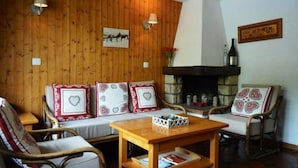 Ein gemütliches Wohnzimmer mit Holzwänden, einem Kamin und einem Steinkaminvorsatz. Zwei Sofas mit gemusterten Kissen und ein Holztisch befinden sich in der Mitte.