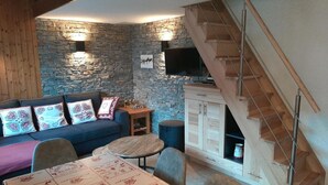 Ein gemütlicher Wohnbereich mit Steinwand, einer Couch mit gemusterten Kissen, einem runden Tisch mit Tischdecke und einer Holztreppe.