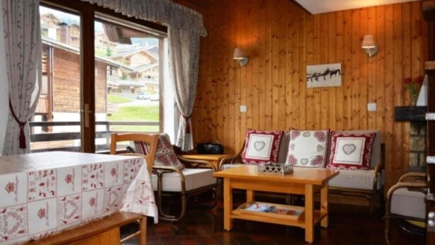 Ein gemütlicher Raum mit Holzwänden, einem Esstisch, einer Couch mit herzförmigen Kissen und einem Fenster mit Vorhängen.
