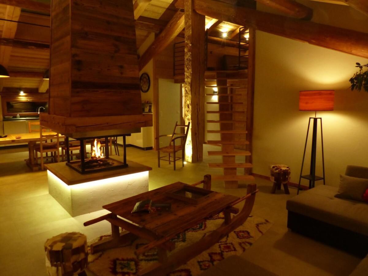 Interior acogedor de una cabaña con chimenea central, muebles de madera y una escalera que lleva a un nivel superior.