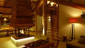 Interior acogedor de una cabaña con chimenea central, muebles de madera y una escalera que lleva a un nivel superior.