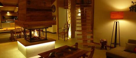 Interior acogedor de una cabaña con chimenea central, muebles de madera y una escalera que lleva a un nivel superior.