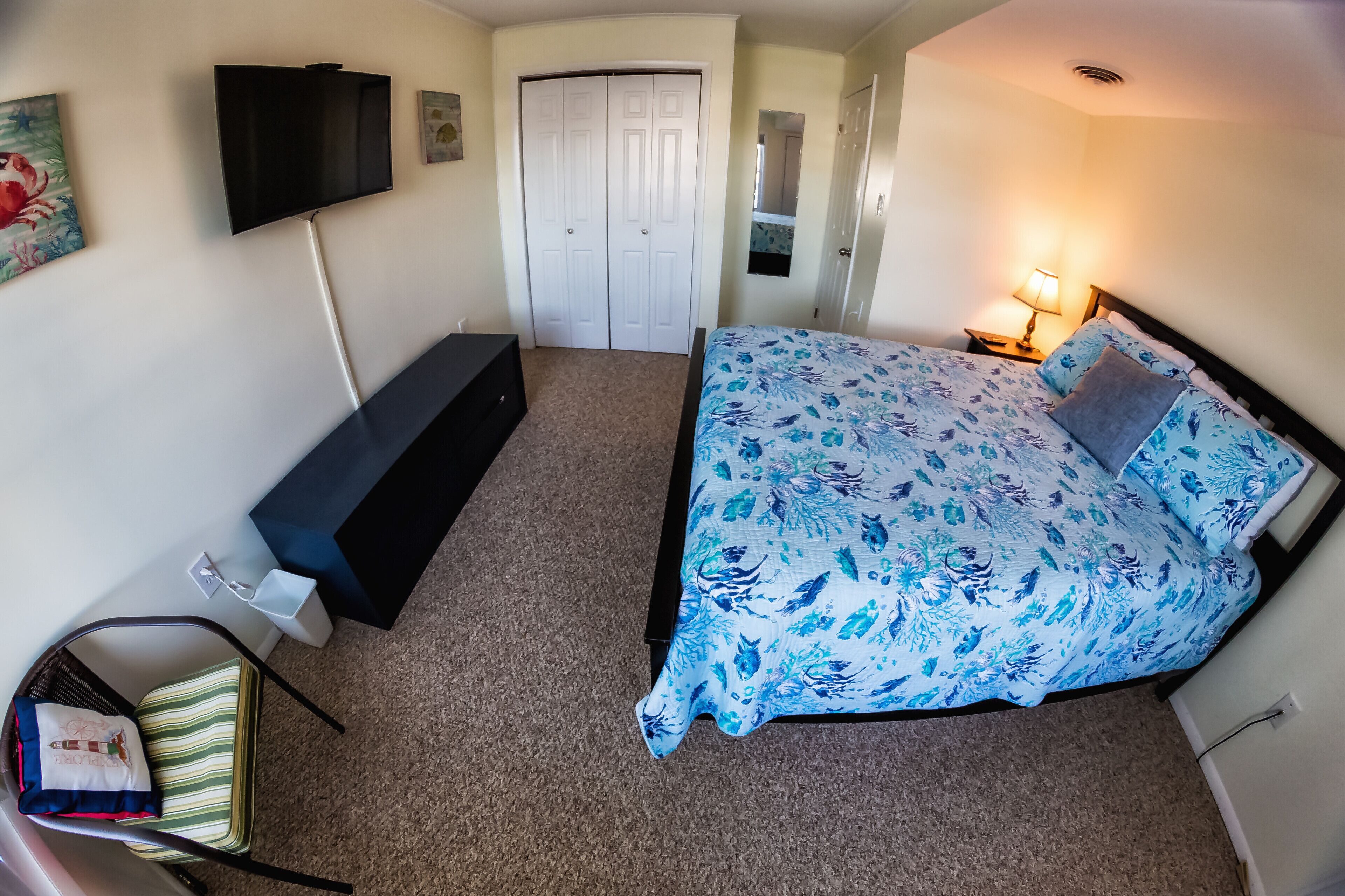 A bedroom with a bed, a TV mounted on the wall, a wardrobe, a chair, and a bedside table with a lamp.