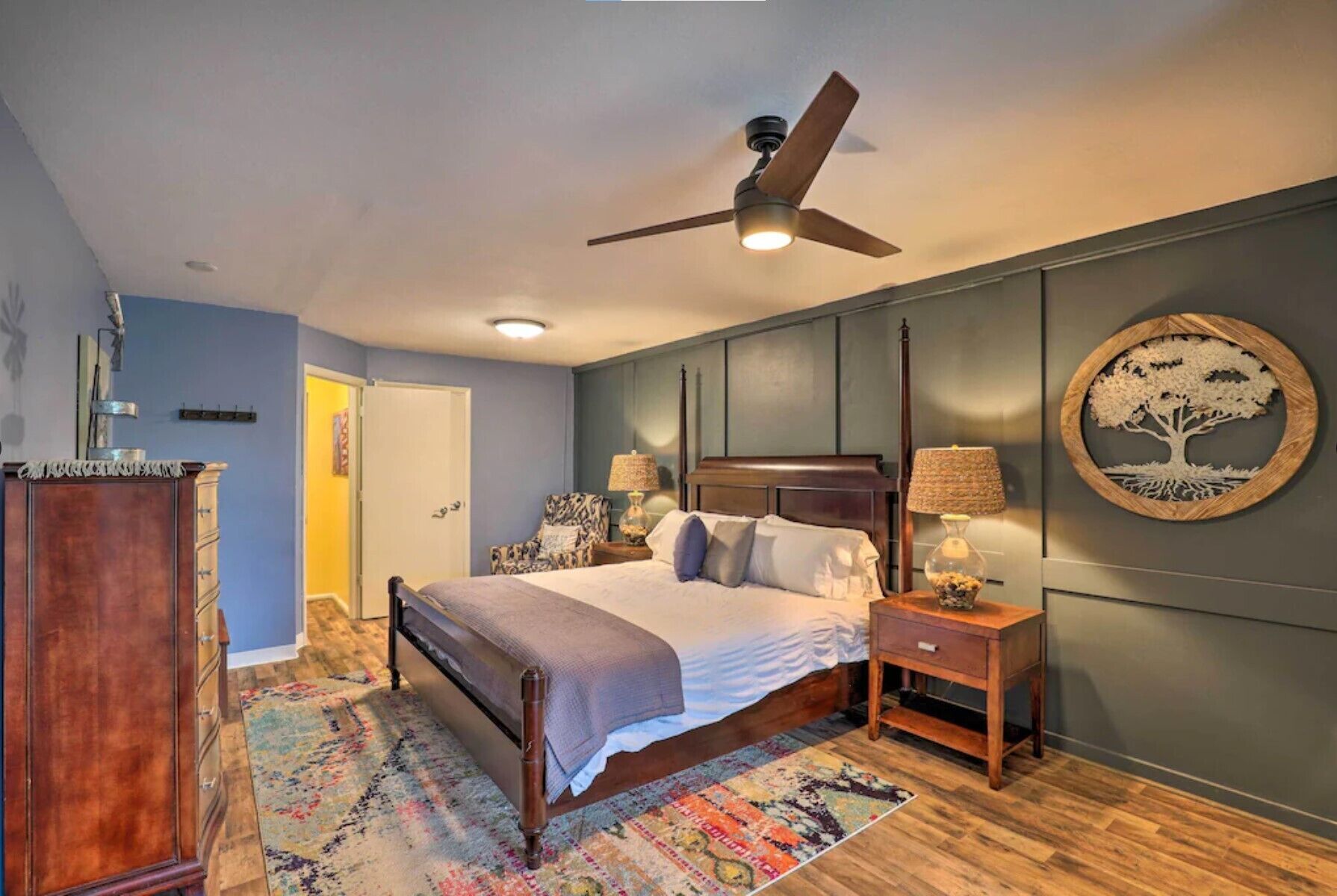 A bedroom with a bed, bedside tables, a ceiling fan, a wall-mounted lamp, and a decorative tree artwork.