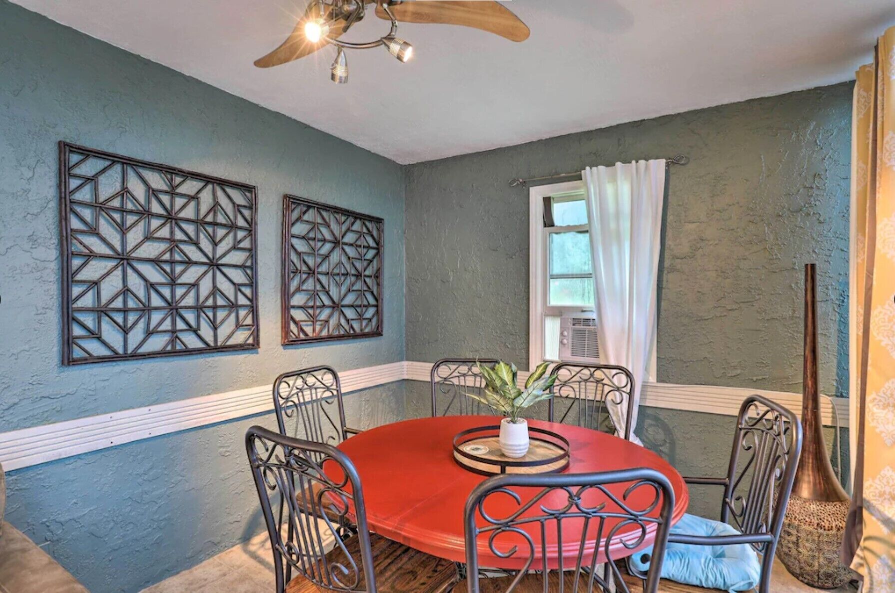 Un comedor con una mesa redonda, sillas de metal, un ventilador de techo, paneles decorativos en la pared y una ventana con cortinas.
