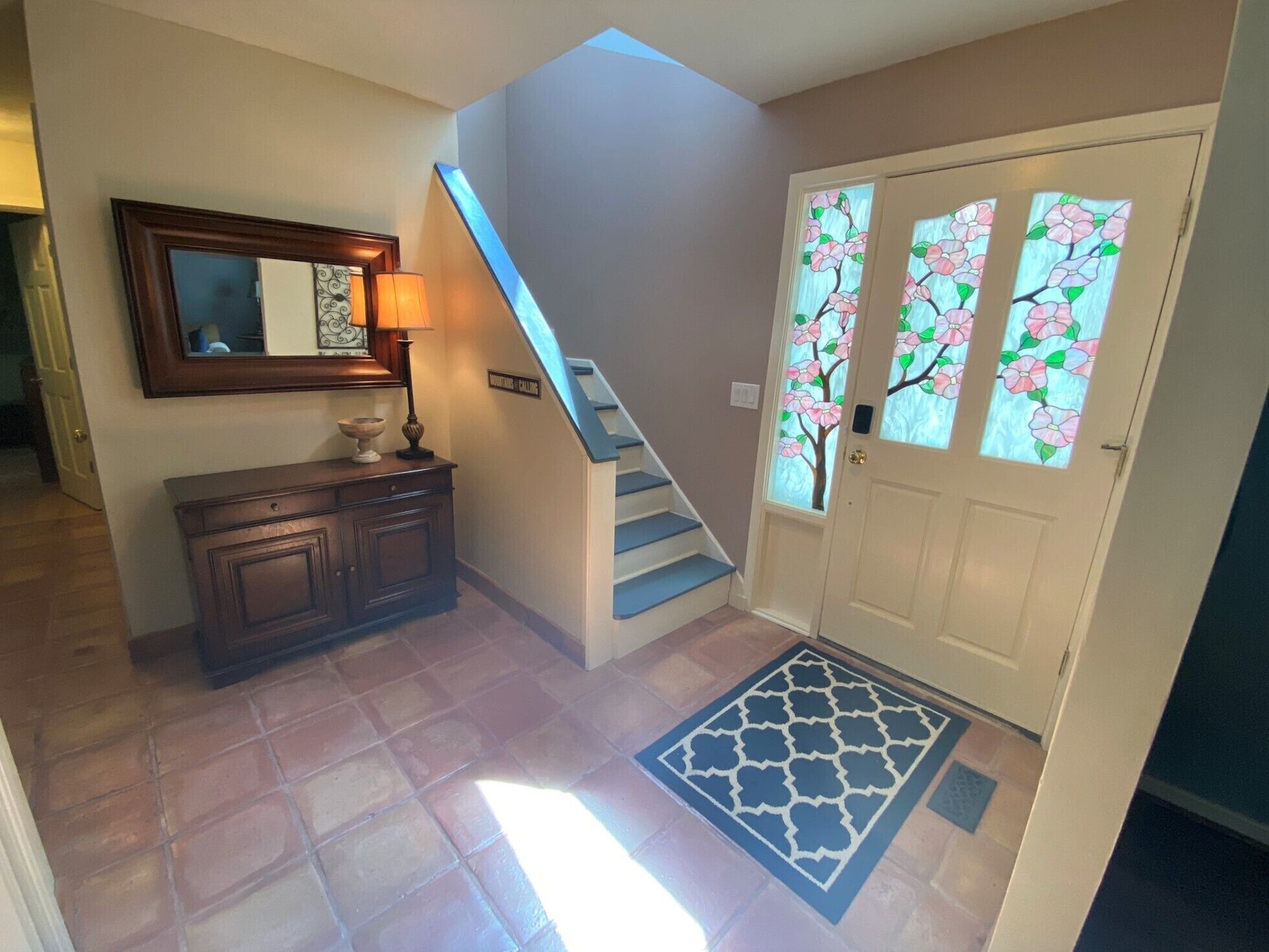 A hallway with a staircase, a door with stained glass, a mirror, and a cabinet.