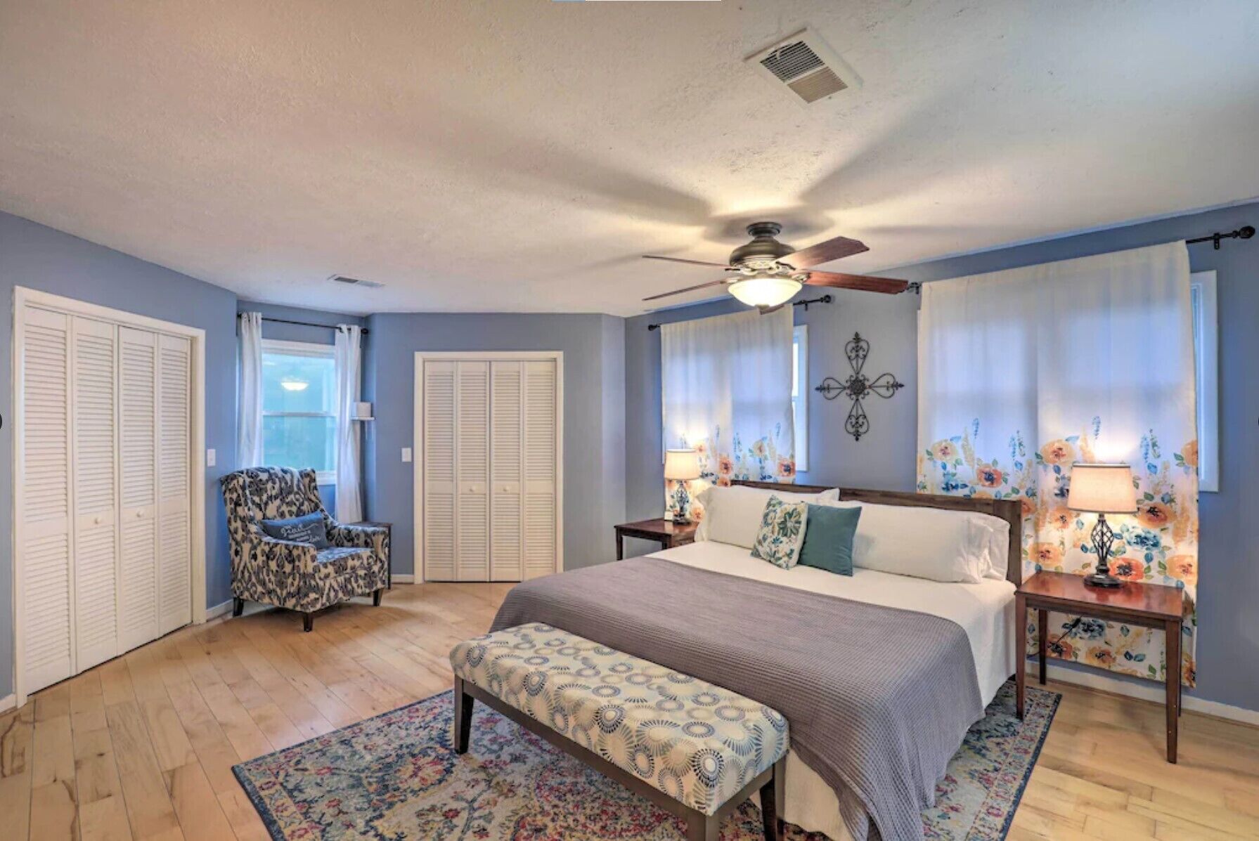 A bedroom with a bed, a chair, a bedside table, a lamp, a ceiling fan, and a window with curtains.