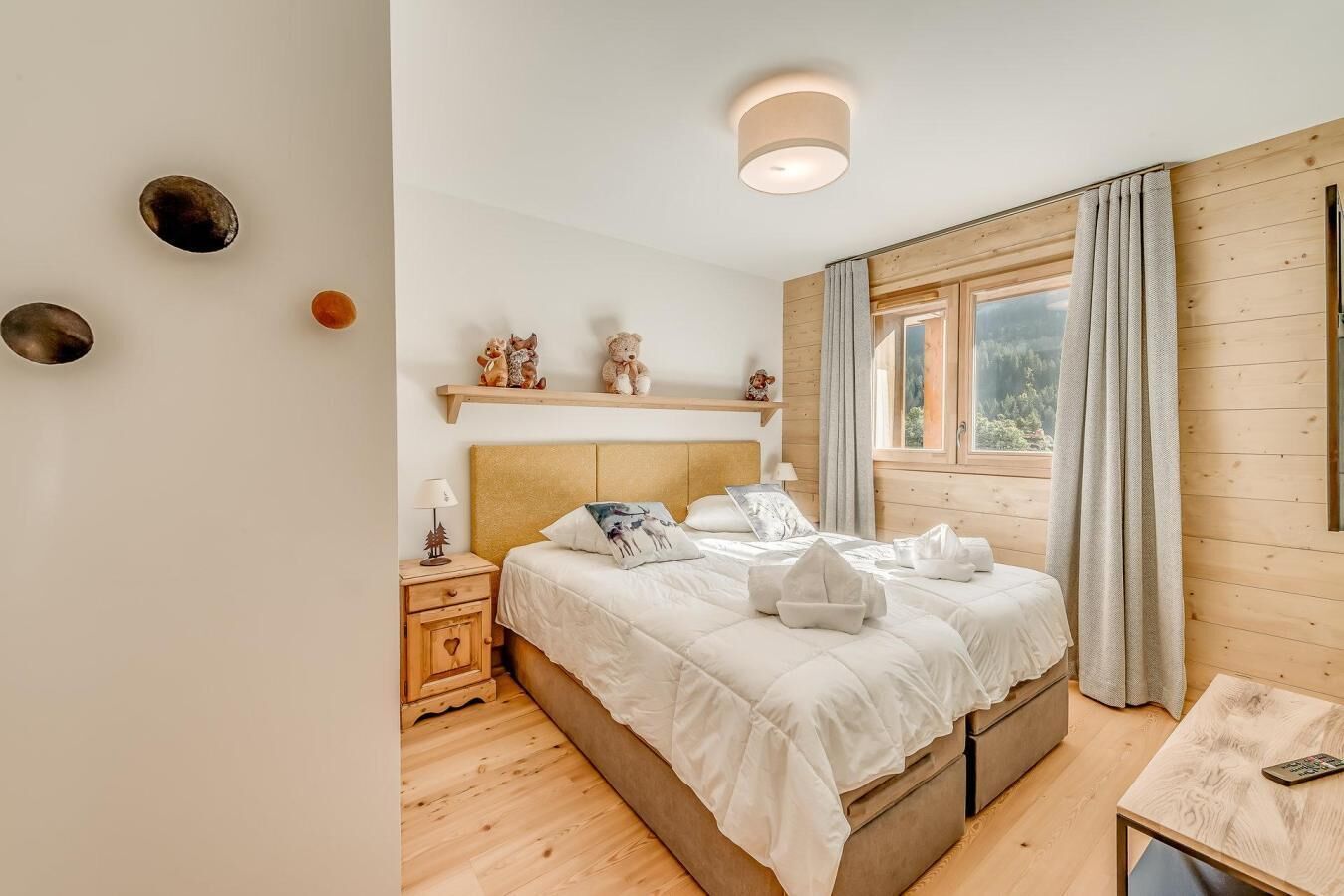A bedroom with a wooden headboard, a bedside table with a lamp, a window with curtains, and a shelf with decorative items.