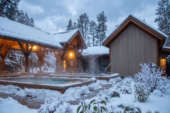 A wooden cabin with a hot tub surrounded by snow, set in a forested area.