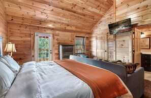 A wooden cabin bedroom with a bed, a sofa, a television mounted on the wall, and a window with blinds. - Luxury Cabin w/ Heated Pool, Theater & Hot Tub | Sleeps 24 (Pigeon Forge)