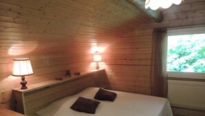 Ein Schlafzimmer einer Holzhütte mit einem Einzelbett, einem Nachttisch, einer Lampe und einem Fenster mit Blick auf Grünflächen.