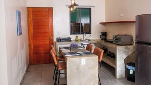 A compact kitchen with a wooden door, a dining table with chairs, a built-in oven, and a refrigerator. - VillasLunaAfricana–101 (Puerto Viejo de Talamanca)