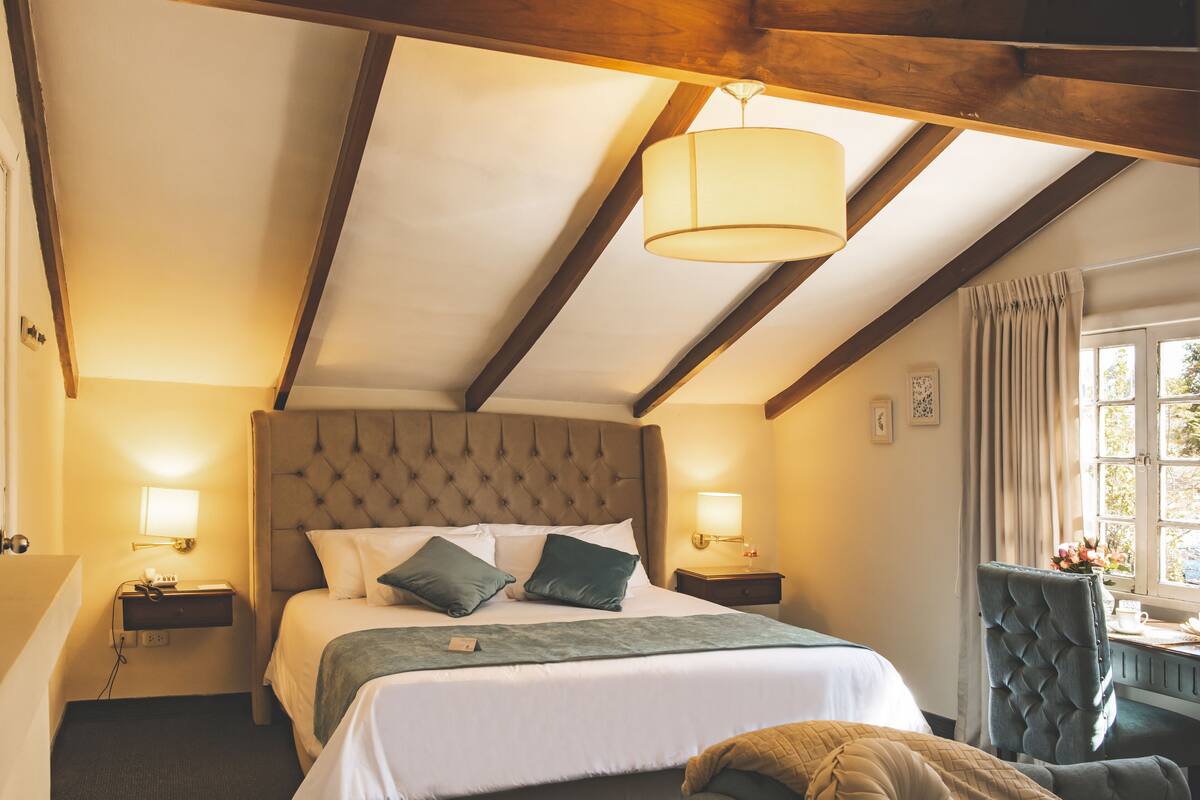 Una habitación de hotel con una cama grande, un cabecero capitonado, dos mesitas de noche con lámparas, una ventana con cortinas y una silla.