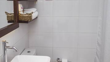 Baño con lavamanos, inodoro y cortina de ducha blancos. Se ve un espejo, un estante con cestas y un bote de basura.