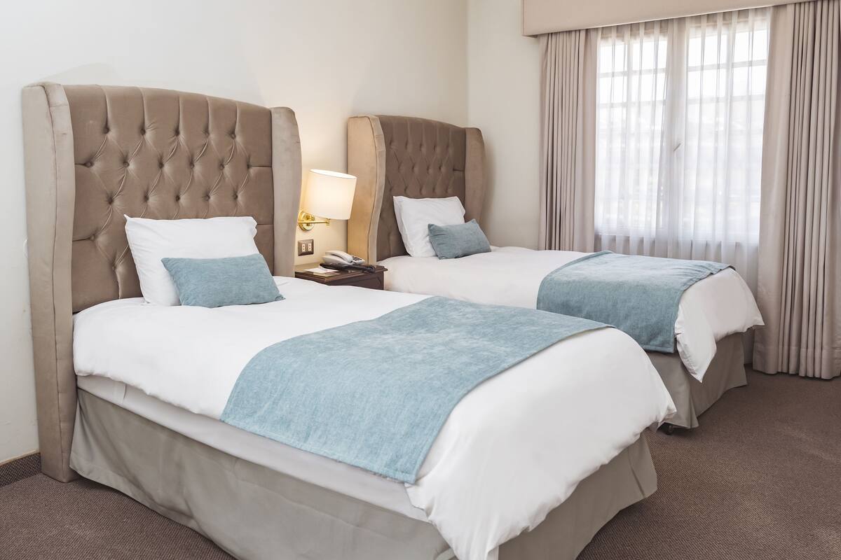 Habitación de hotel con dos camas, cada una con cabecero tapizado, ropa de cama blanca y una manta azul. Hay una mesita de noche con una lámpara y un teléfono.