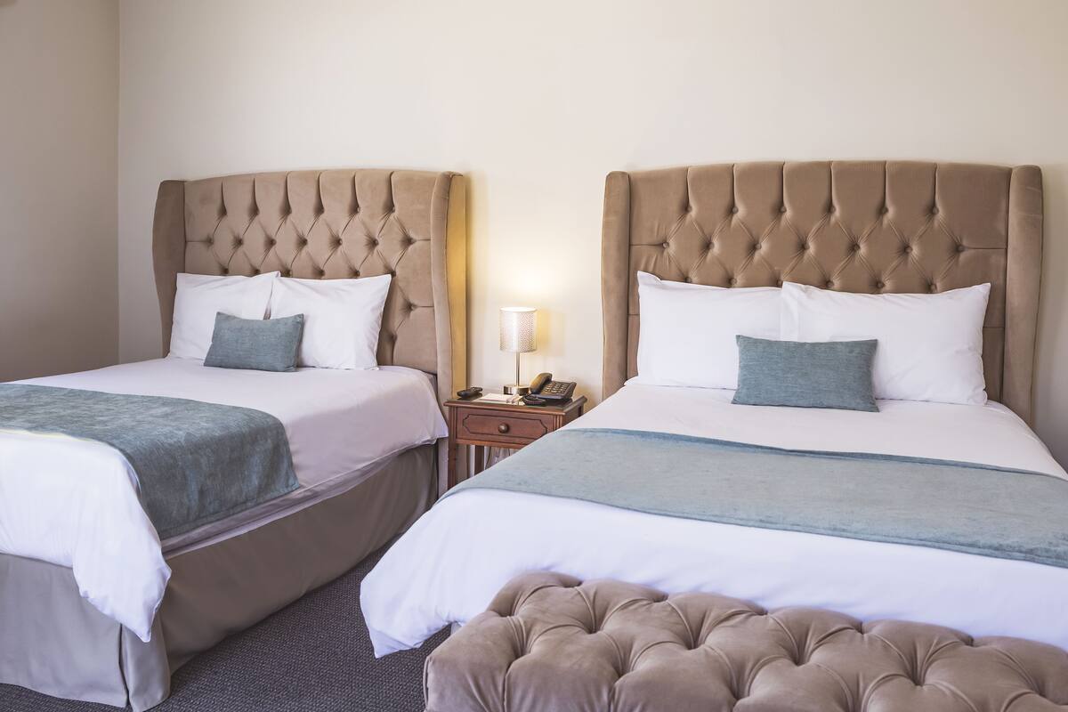 Una habitación de hotel con dos camas, cada una con un cabecero capitonado, ropa de cama blanca y una manta gris. Hay una mesita de noche con una lámpara y un teléfono.