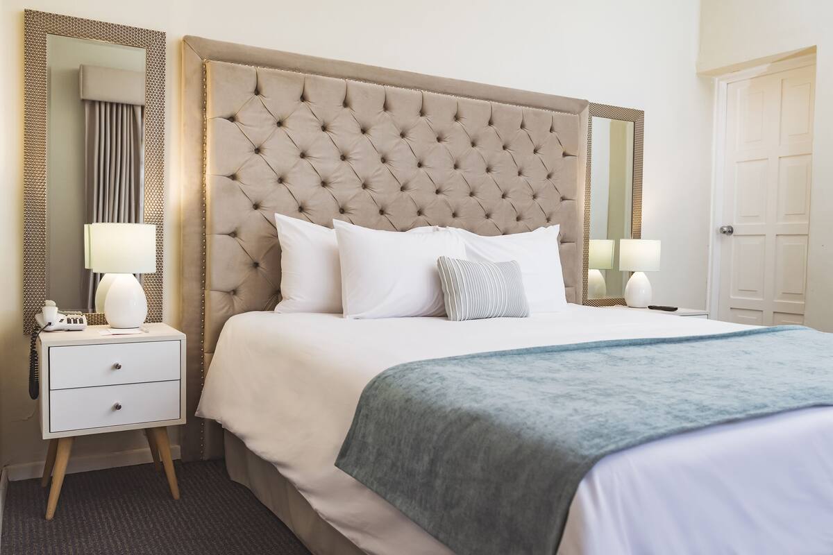 Una cama bien hecha con cabecera acolchada, ropa de cama blanca y una manta azul. Se aprecia una mesita de noche con dos lámparas y un teléfono.