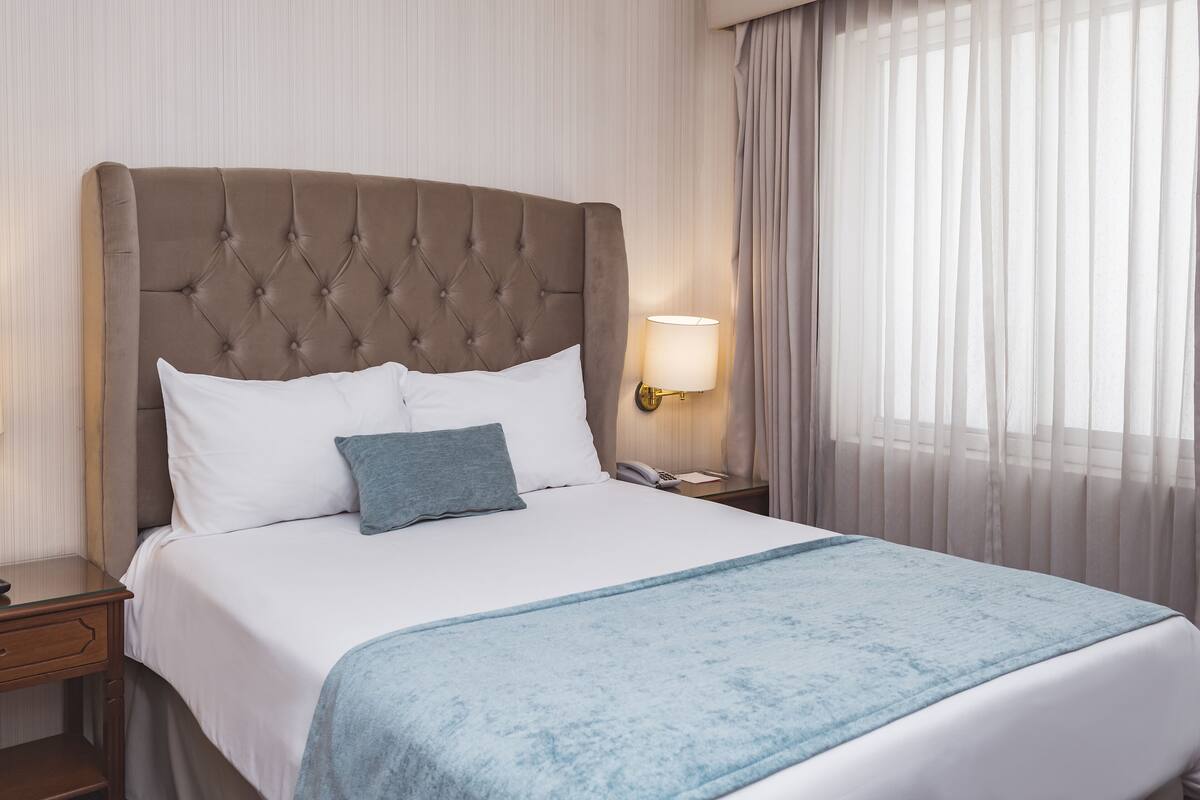 Una cama bien tendida con un cabecero capitonado, ropa de cama blanca y una manta azul.