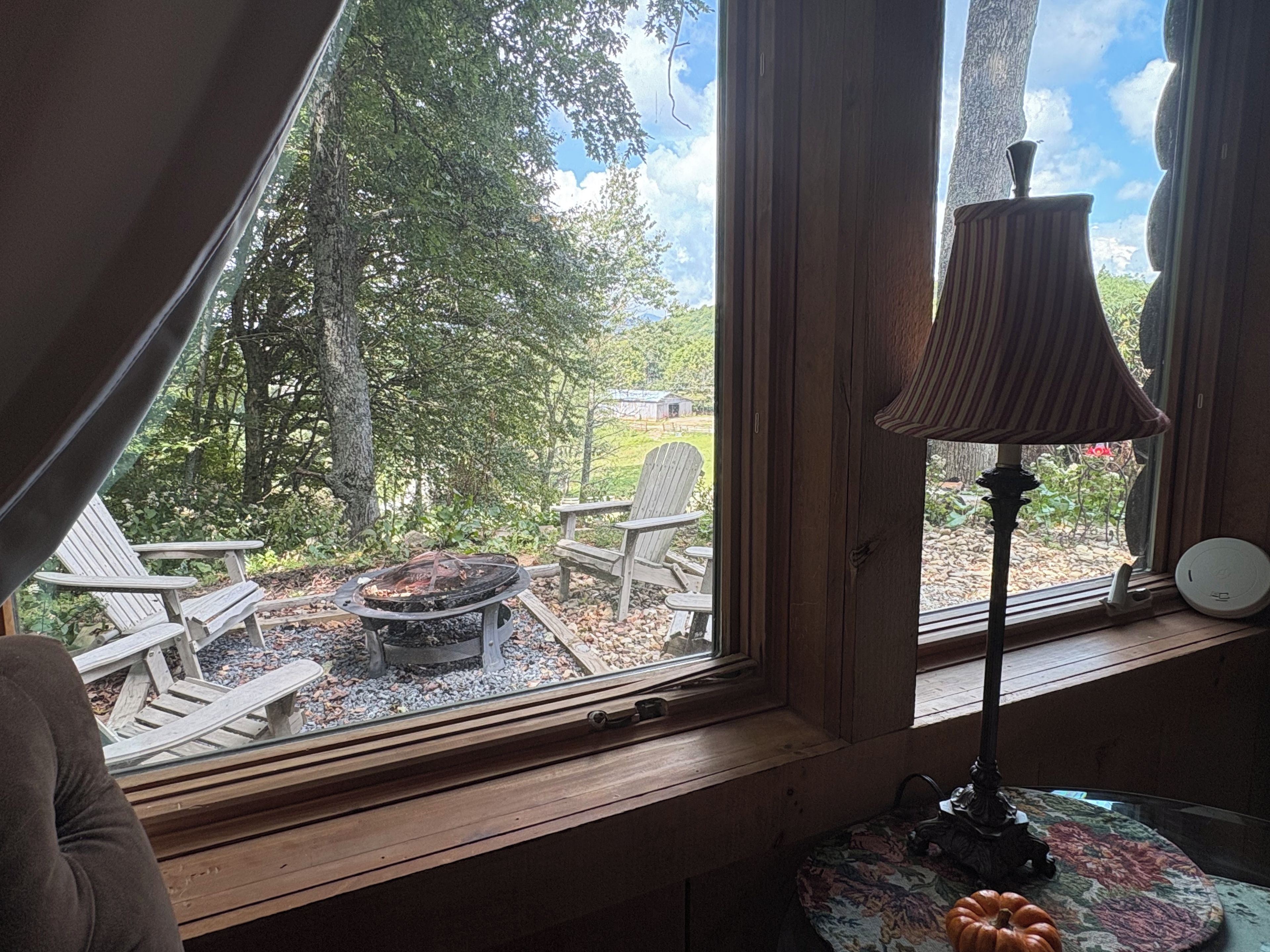 Una habitación con ventana que da a un jardín, un banco de madera, una mesa con lámpara y una pequeña calabaza.