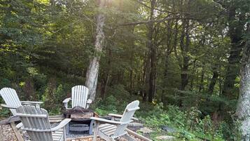 A forested area with a fire pit and Adirondack chairs.