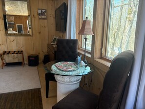 Une pièce chaleureuse aux murs en bois, avec une table en verre ornée d’une assiette décorative, un fauteuil rembourré, une lampe et une fenêtre donnant sur des arbres.