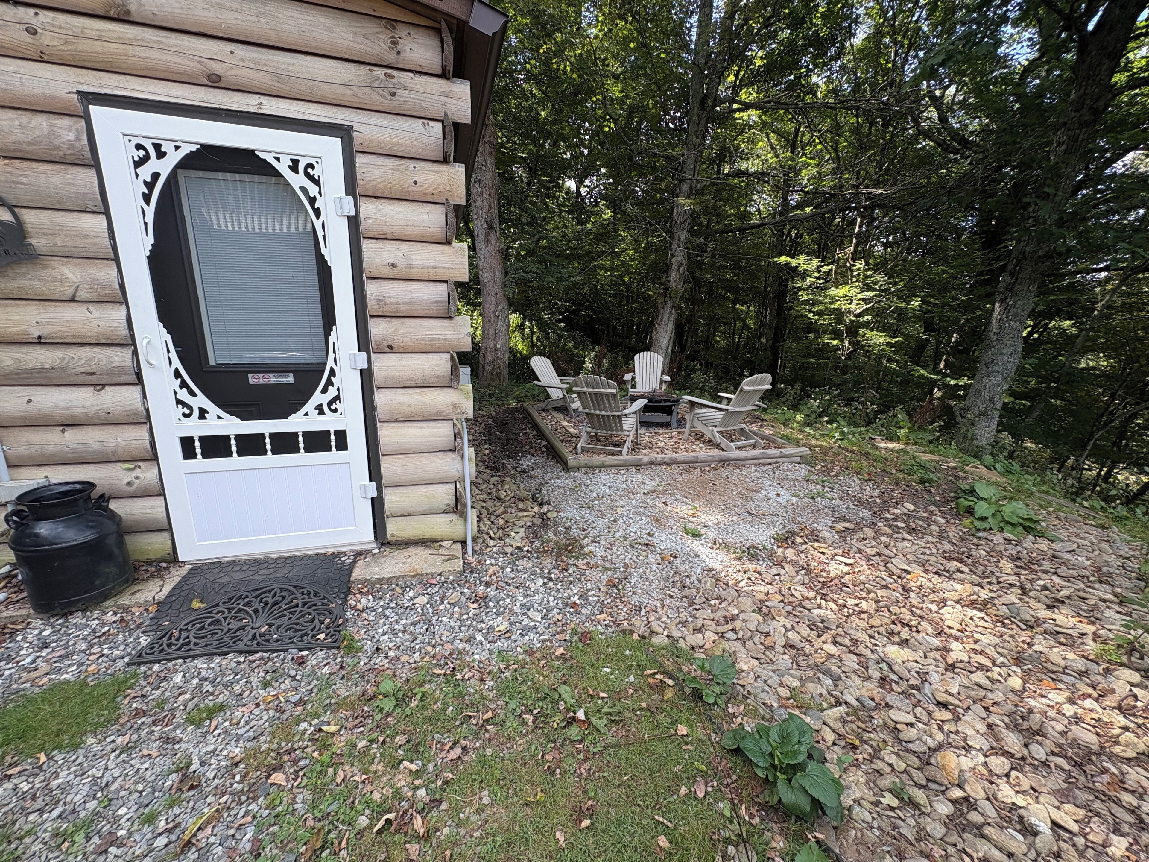 A log cabin with a white door, a fire pit, and Adirondack chairs in a wooded area.
