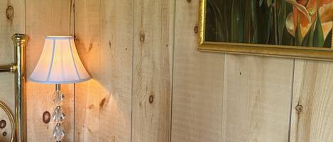 A wooden wall with a framed painting of flowers, a table with a lamp and a pumpkin, and a brass lamp on the left.