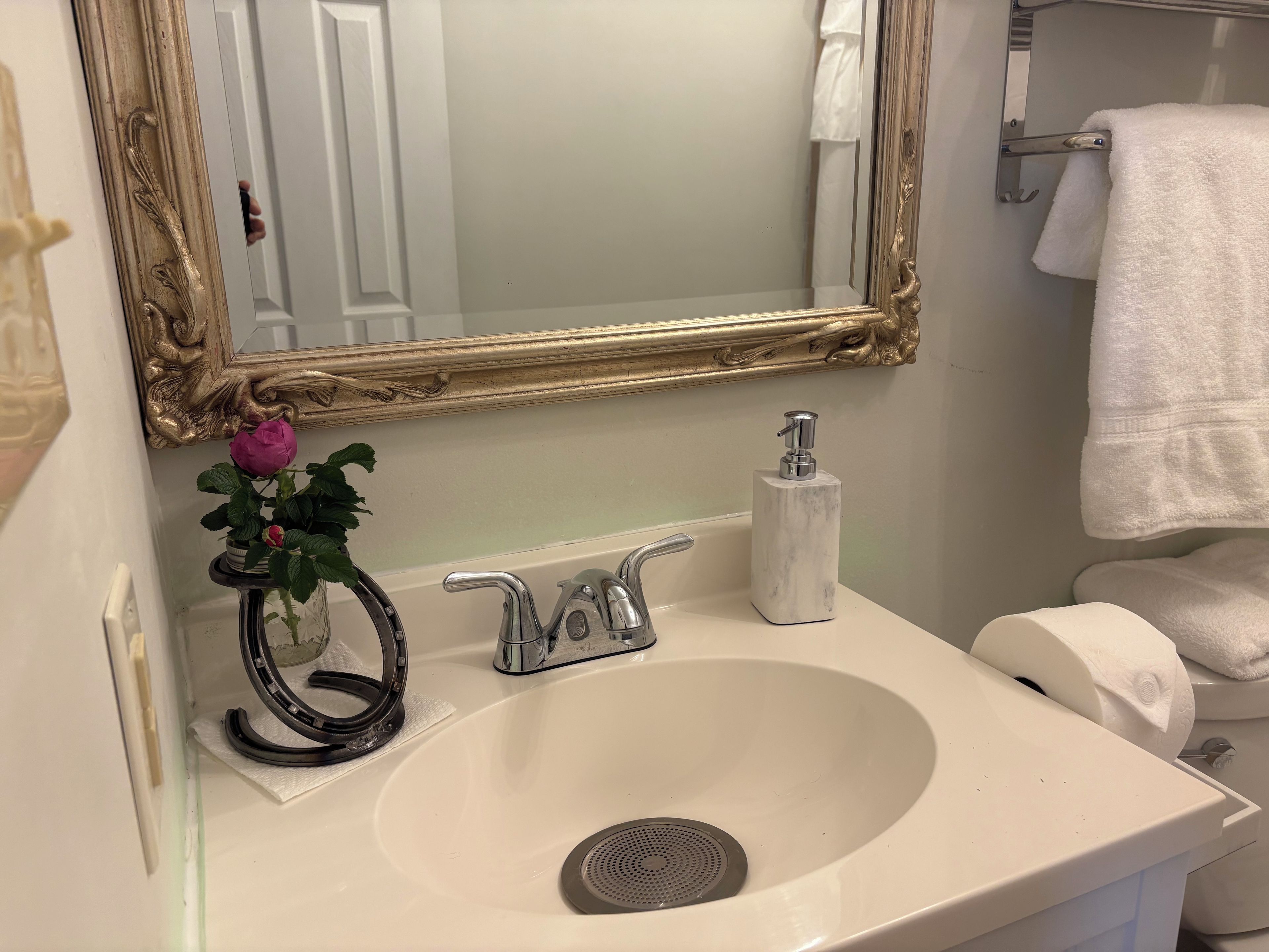 Un baño con un lavamanos blanco, un espejo con marco dorado, una rosa rosa y una toalla blanca.
