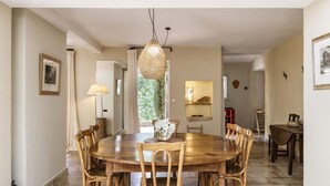 A dining area with a round wooden table, six wooden chairs, a hanging light fixture, and a view of a garden through a window. - Pool, Villa La Colline, beautiful view of Mt Ventoux (Suzette)