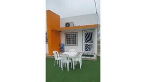 Un bâtiment à la façade blanche et orange, doté d’un espace extérieur abrité avec des chaises en plastique blanc et une table, ainsi que d’une pelouse artificielle verte.